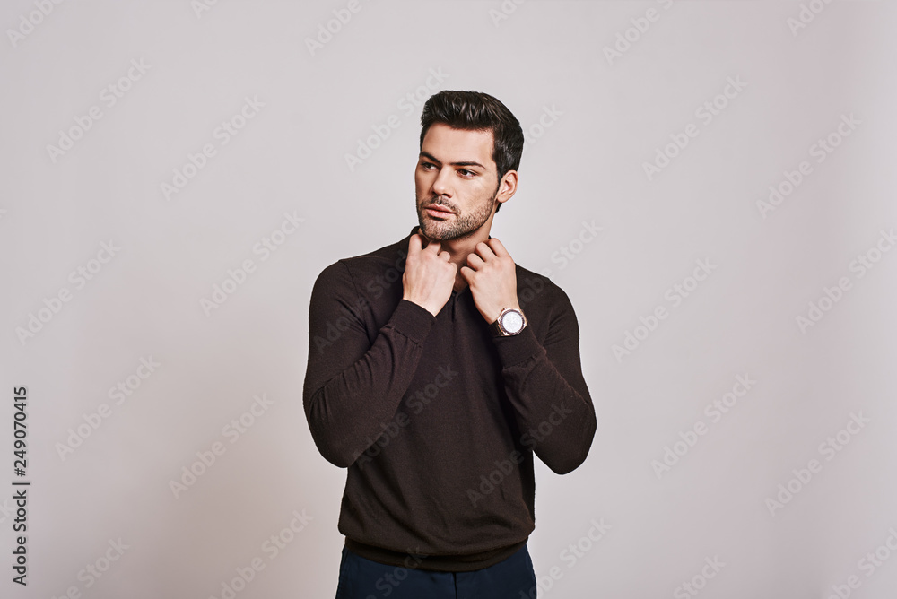 He cares about his style. Young dark-haired man wearing brown jumper standing over grey background