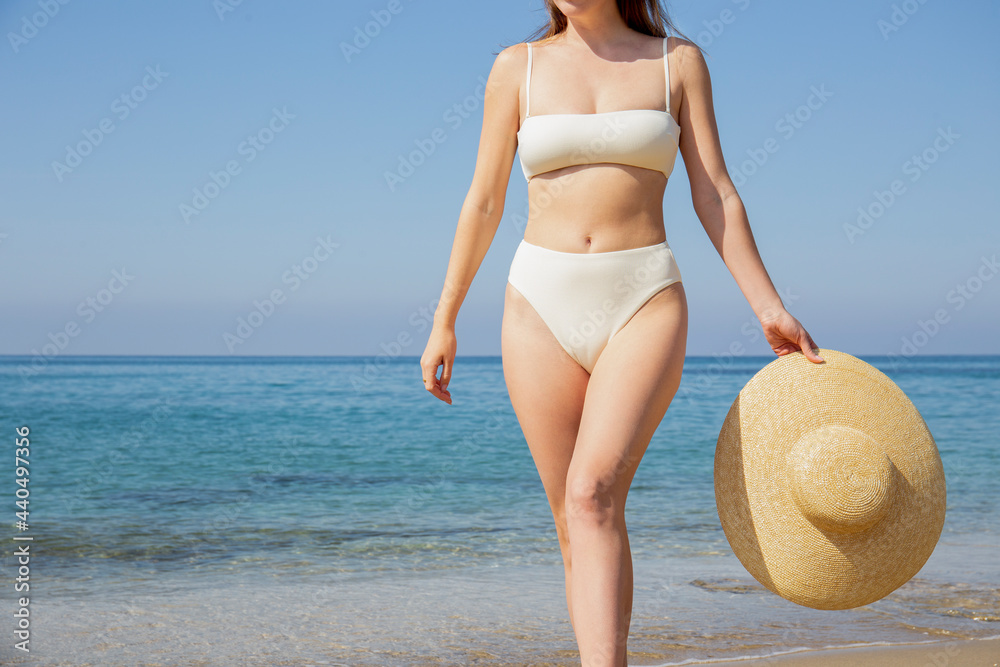 Caucasian woman with bikini bathing suit with high waisted bottom bikini and broad brim straw hat posing at sandy beach on beautiful sunny day.