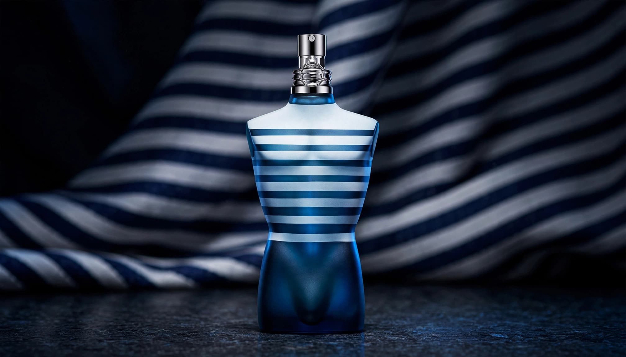 A torso-shaped fragrance bottle on a dark counter with blue sailor-inspired stripes behind it.