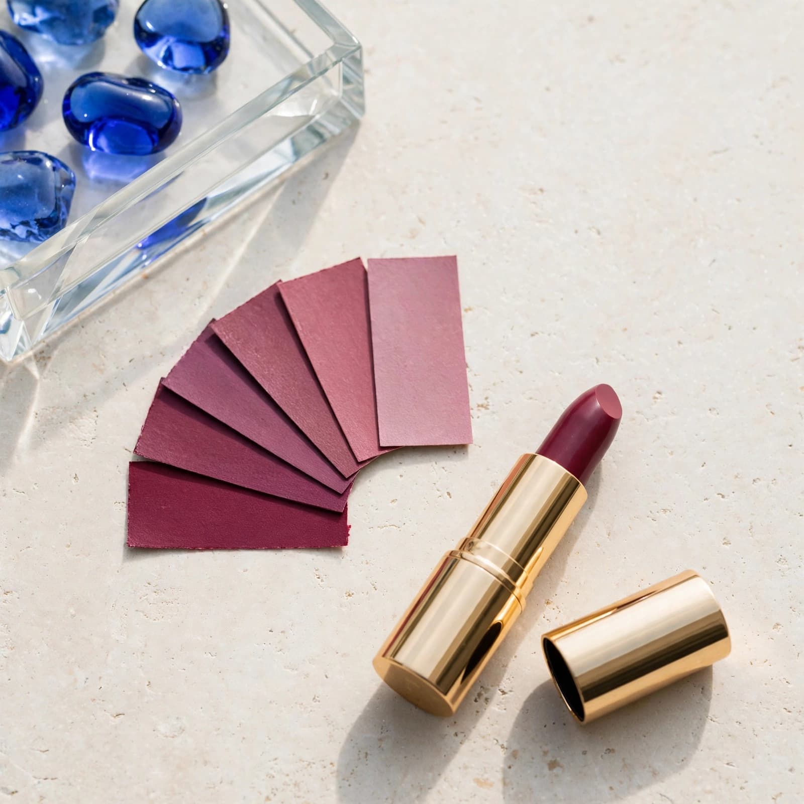 A berry lipstick and colour swatches sit on a pale stone table beside a blue glass tray.