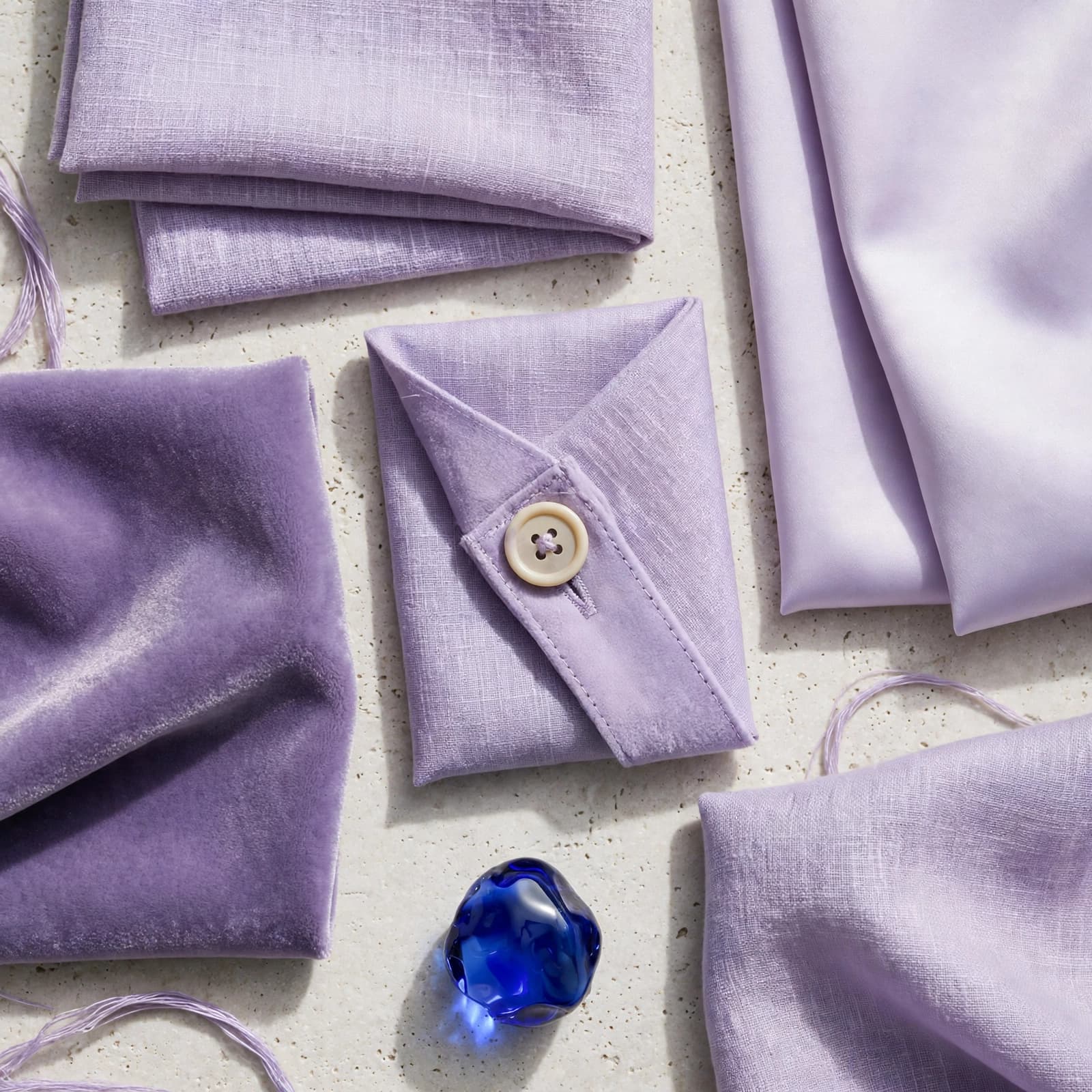 Lavender dress fabrics show different textures, a seam, a button, and a blue glass accent.